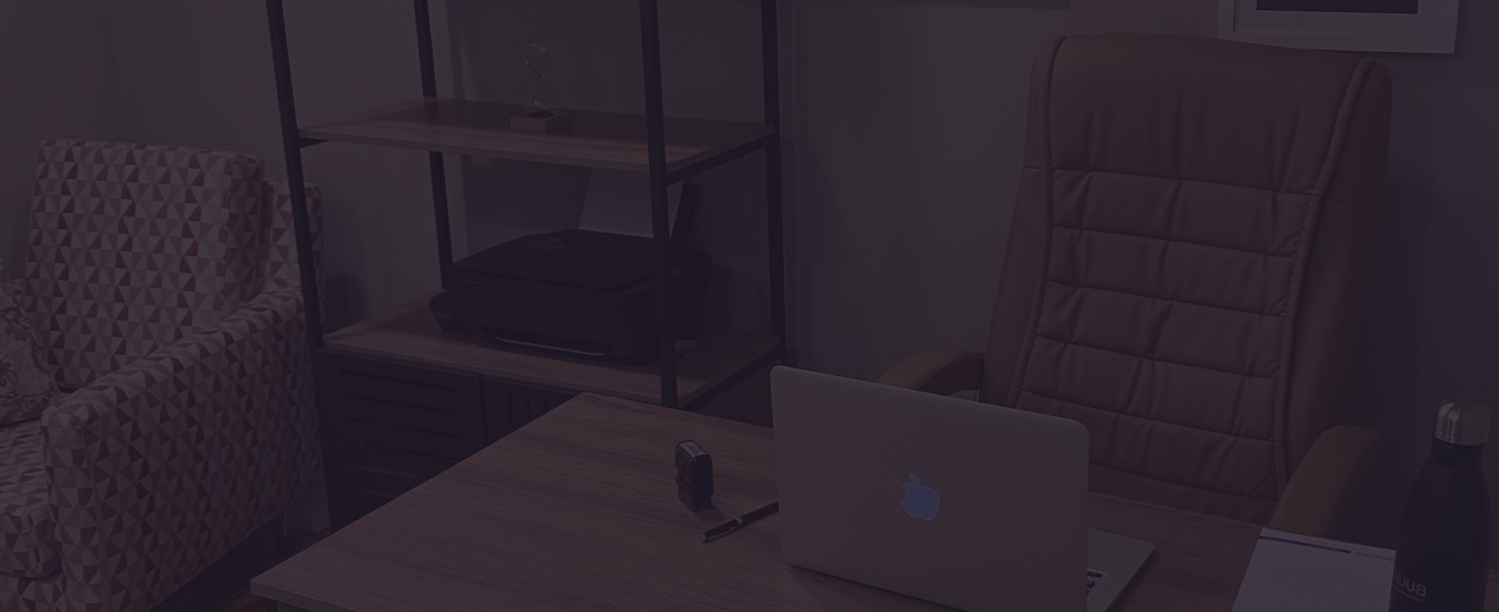 Sala de um consultório para atendimento à saúde mental. O ambiente tem três poltronas. Em cima da mesa tem um notebook, garrafa e papéis. Também há uma prateleira.