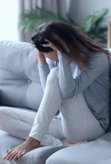 Mulher sentada em um sofá com umas das pernas sobre o móvel. Ela está com os cabelos soltos e cabeça baixa. As suas duas mãos estão sobre o início da sua cabeça.