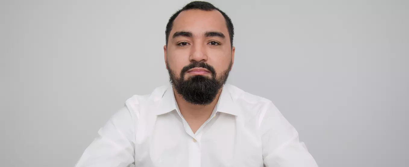 Homem sentado em frente a uma mesa mexendo em um notebook. Ele usa uma camisa social e um relógio em seu pulso.